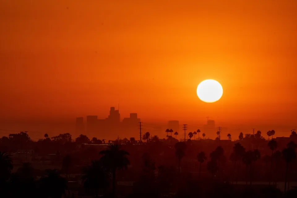 Sun rising over downtown Los Angeles with tall palm trees visible in foreground. Image is bright orange-red indicating extreme heat.
